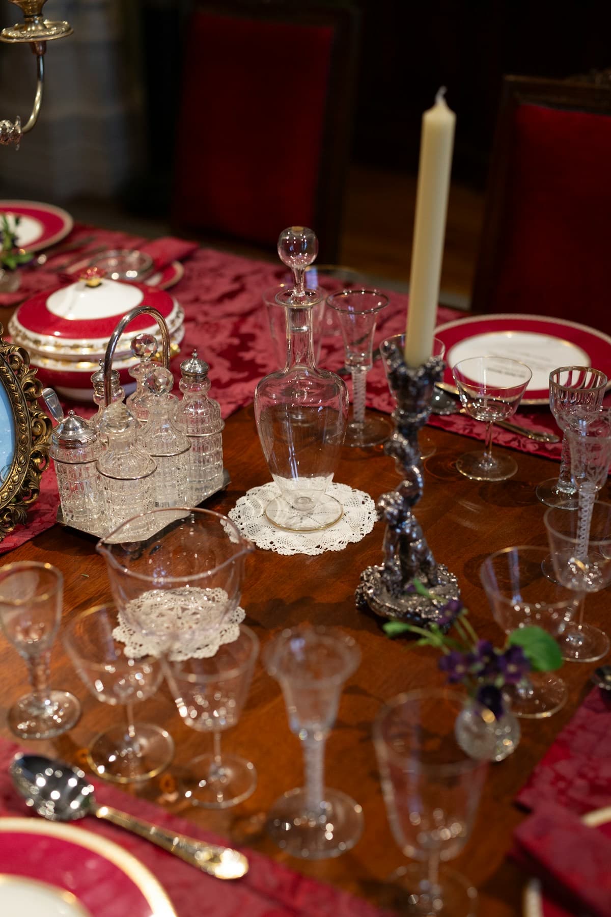 A laid table of antique crystal stemware, a cut-glass cruet set, a silver candlestick and gilt-rimmed crimson china on a damask runner.