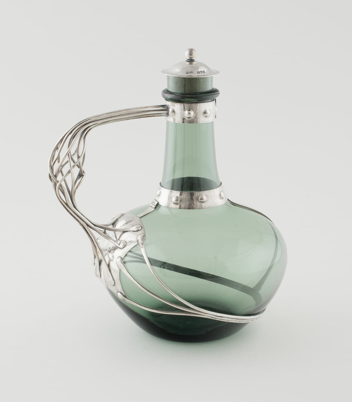 A green glass decanter with a hammered silver collar, lid and looping silver handle.
