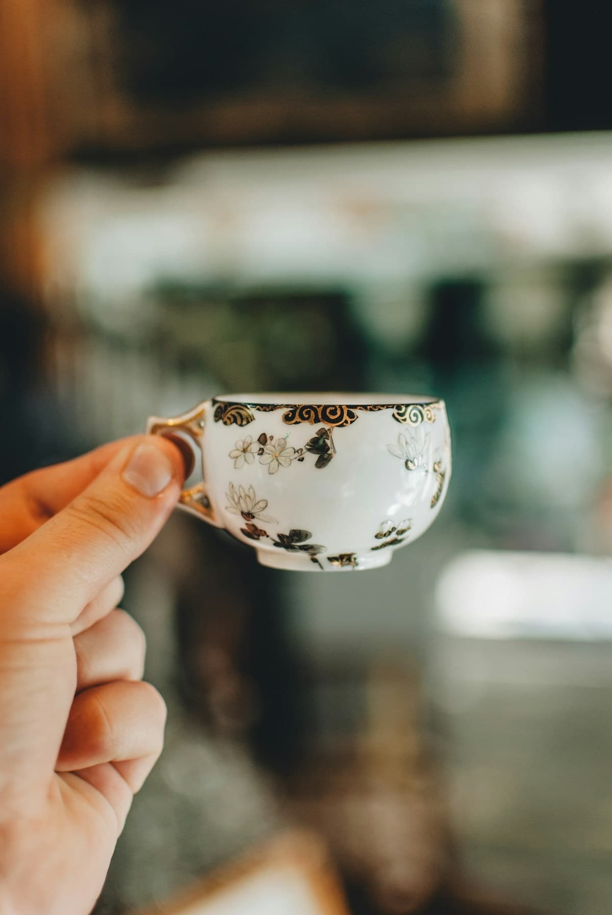 A hand holding a small gilt-decorated porcelain teacup painted with white blossoms.